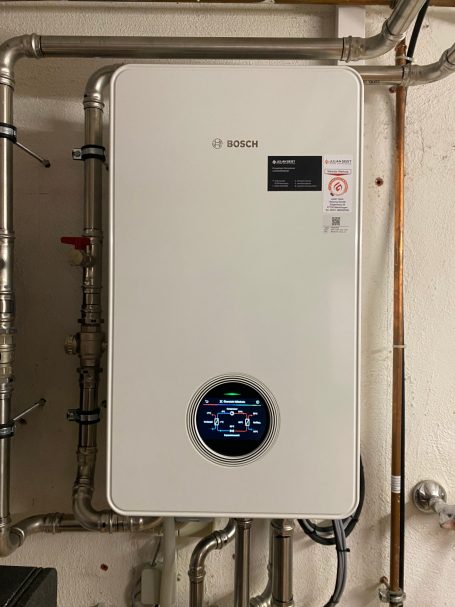 Weiße Heizungsanlage an der Wand mit digitalem Display und Rohrleitungen.